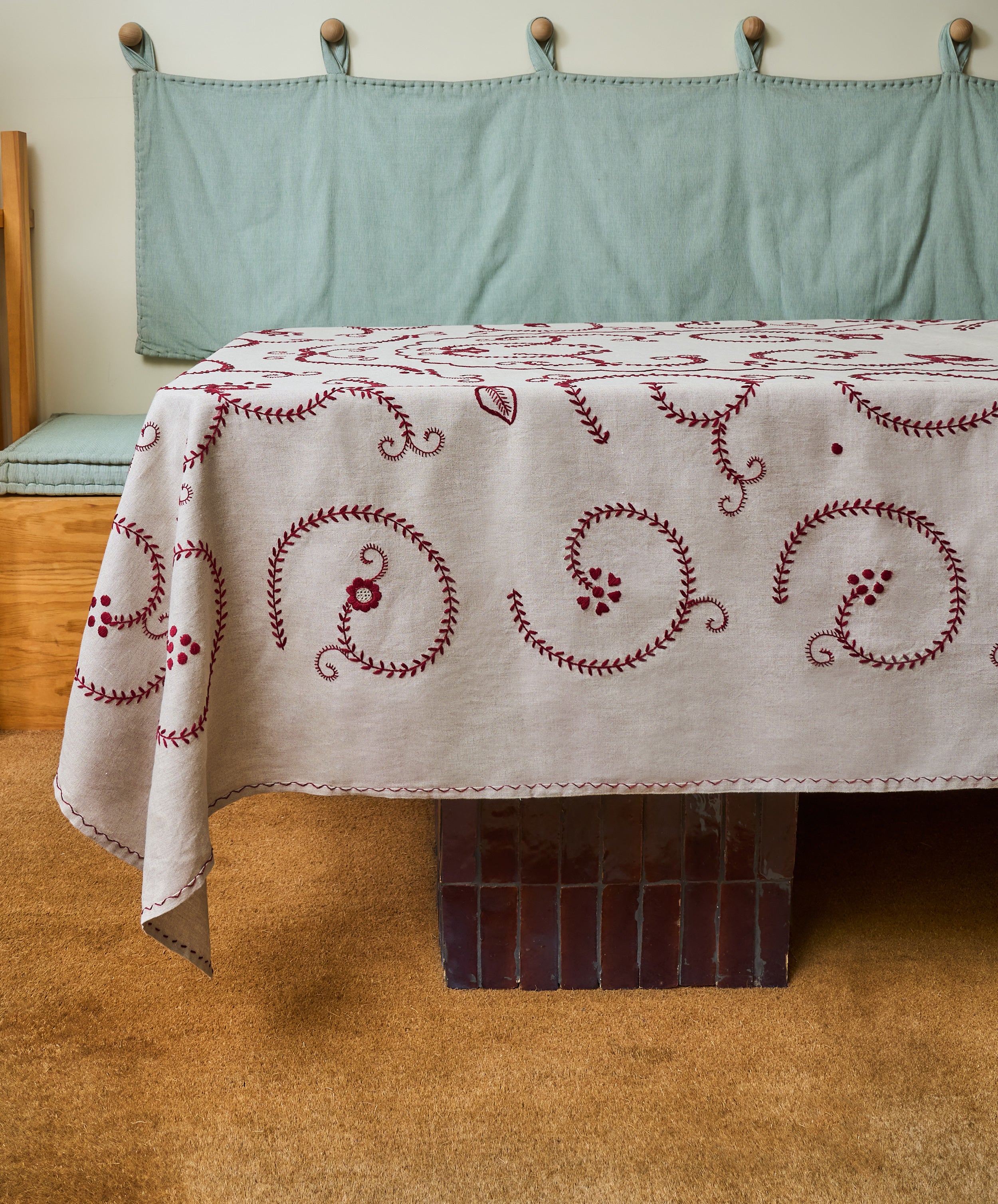The Inez Tablecloth, crafted from raw linen with hand-embroidered red details, sits atop a brown-tiled table. A bench with a blue cushion completes the scene, showcasing traditional Portuguese design.