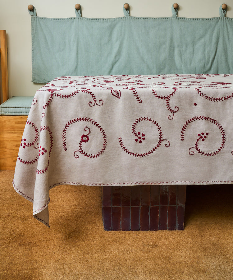 The Inez Tablecloth, crafted from raw linen with hand-embroidered red details, sits atop a brown-tiled table. A bench with a blue cushion completes the scene, showcasing traditional Portuguese design.