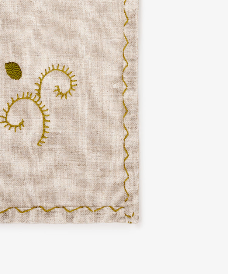 A close-up of the Inez Napkin, Set of 6, showcasing raw linen fabric with green hand-embroidered patterns and a scalloped green stitched border—an elegant touch for your table setting.