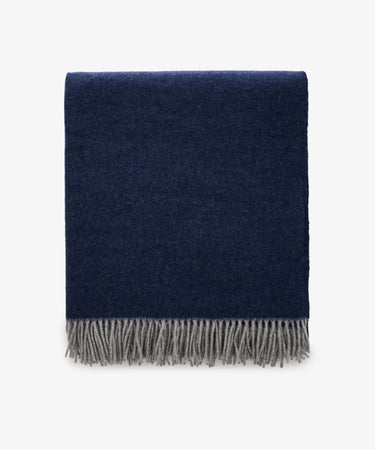The Ulisse Throw, in oceanic blue with a gray fringe, is neatly folded and displayed against a plain white background.