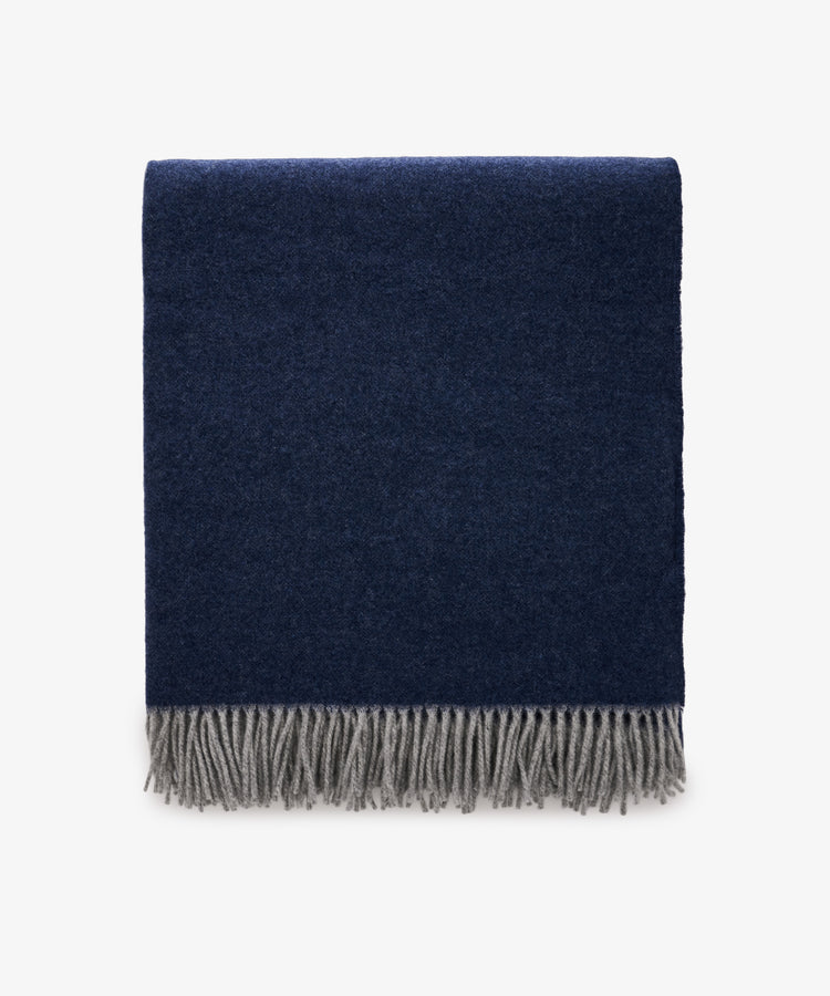 The Ulisse Throw, in oceanic blue with a gray fringe, is neatly folded and displayed against a plain white background.