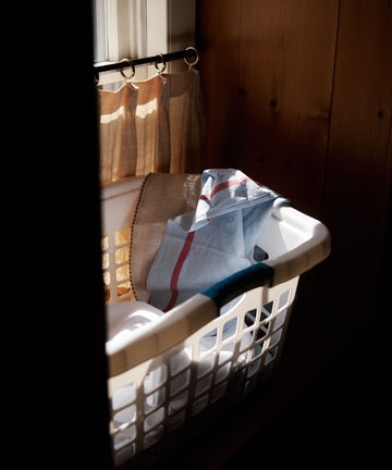 A white laundry basket filled with folded Métis Tea Towels sits by a window with sheer curtains, illuminated by natural sunlight.