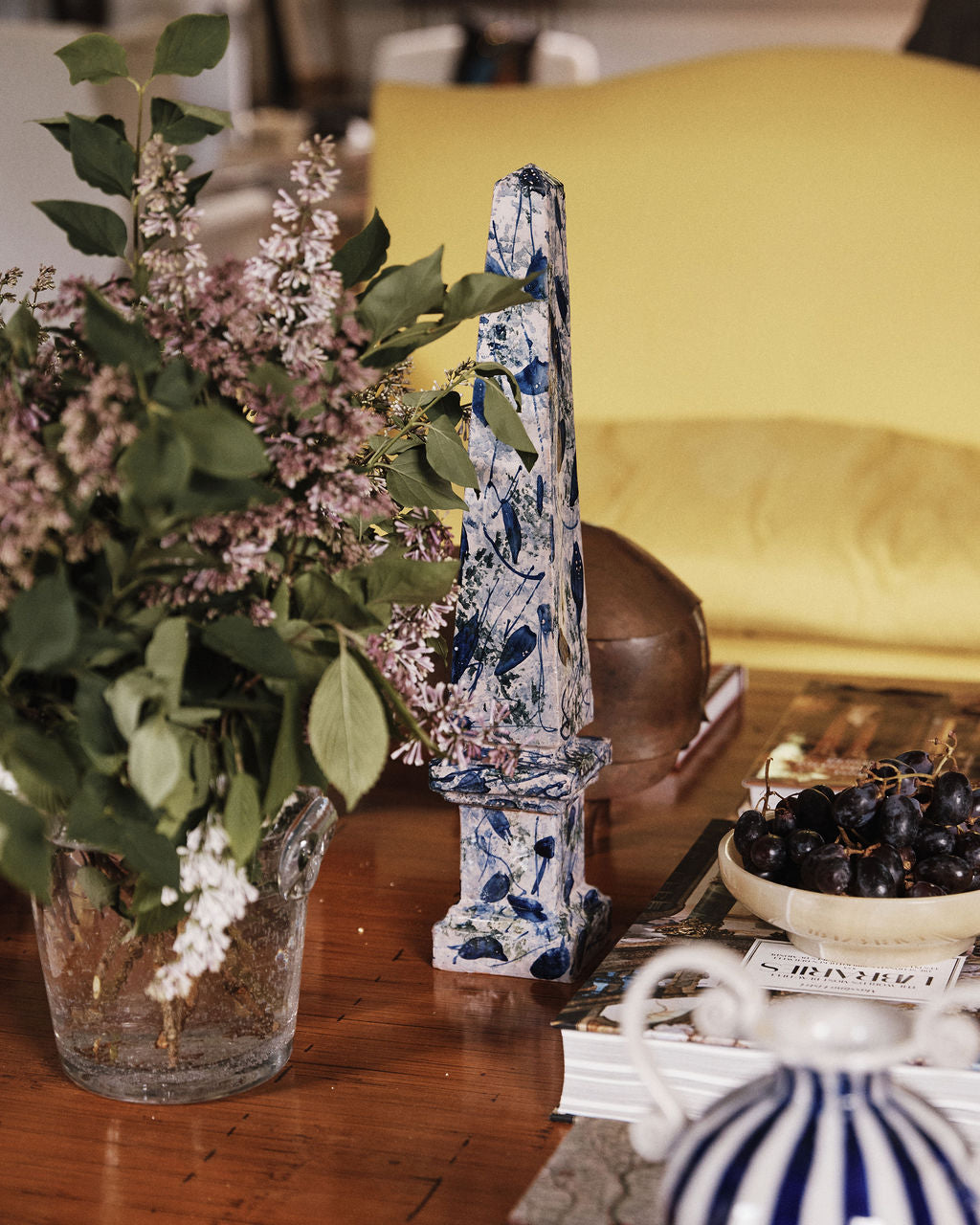 A glass vase of lilacs, the Armando Obelisque, a bowl of grapes, and stacked books on a wooden table in front of a yellow sofa evoke the warmth of Seville.