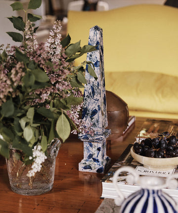 A glass vase of lilacs, the Armando Obelisque, a bowl of grapes, and stacked books on a wooden table in front of a yellow sofa evoke the warmth of Seville.