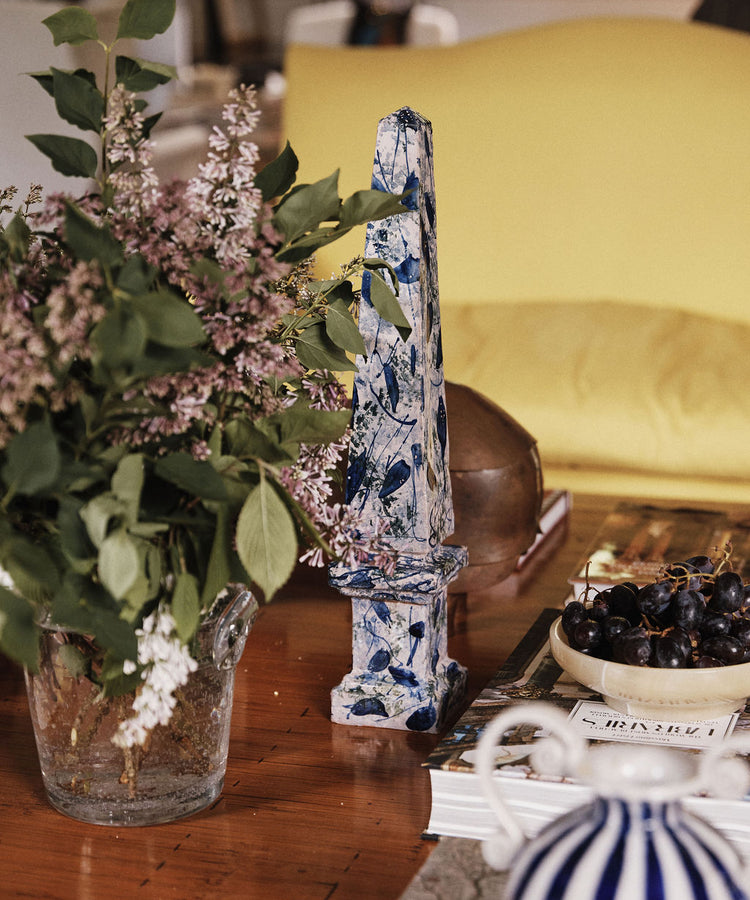 A glass vase of lilacs, the Armando Obelisque, a bowl of grapes, and stacked books on a wooden table in front of a yellow sofa evoke the warmth of Seville.