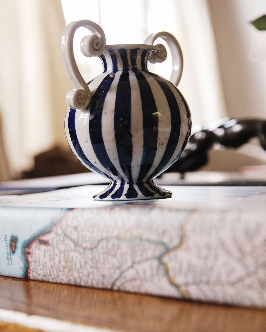 The Cyrene Bud Vase, featuring a hand-thrown design with vertical blue-and-white stripes, sits atop a closed book adorned with a map-themed cover.
