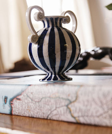 The Cyrene Bud Vase, featuring a hand-thrown design with vertical blue-and-white stripes, sits atop a closed book adorned with a map-themed cover.
