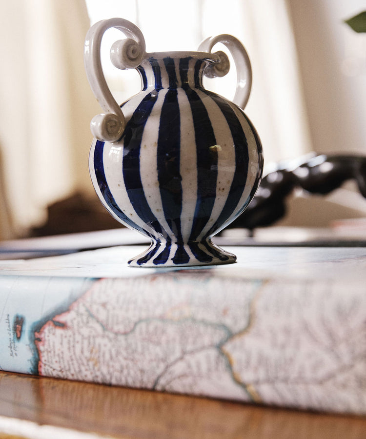The Cyrene Bud Vase, featuring a hand-thrown design with vertical blue-and-white stripes, sits atop a closed book adorned with a map-themed cover.
