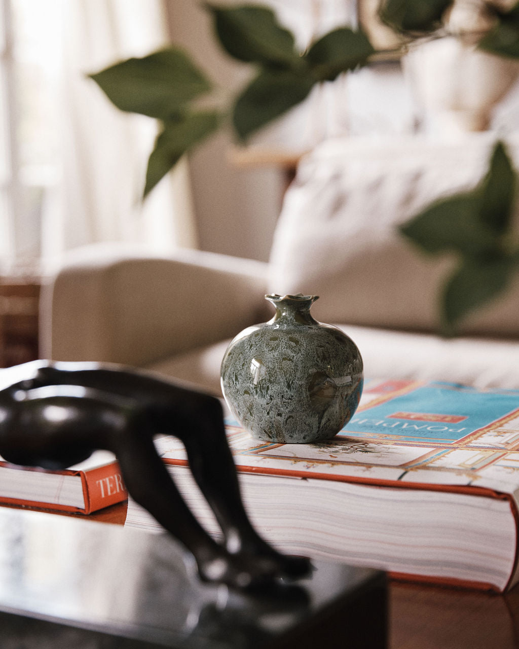 A Pom Bud Vase in green, inspired by handmade ceramic pomegranate designs from Granada ceramics, sits atop two stacked books on a table, with a blurred black sculpture in the foreground and a beige couch in the background.