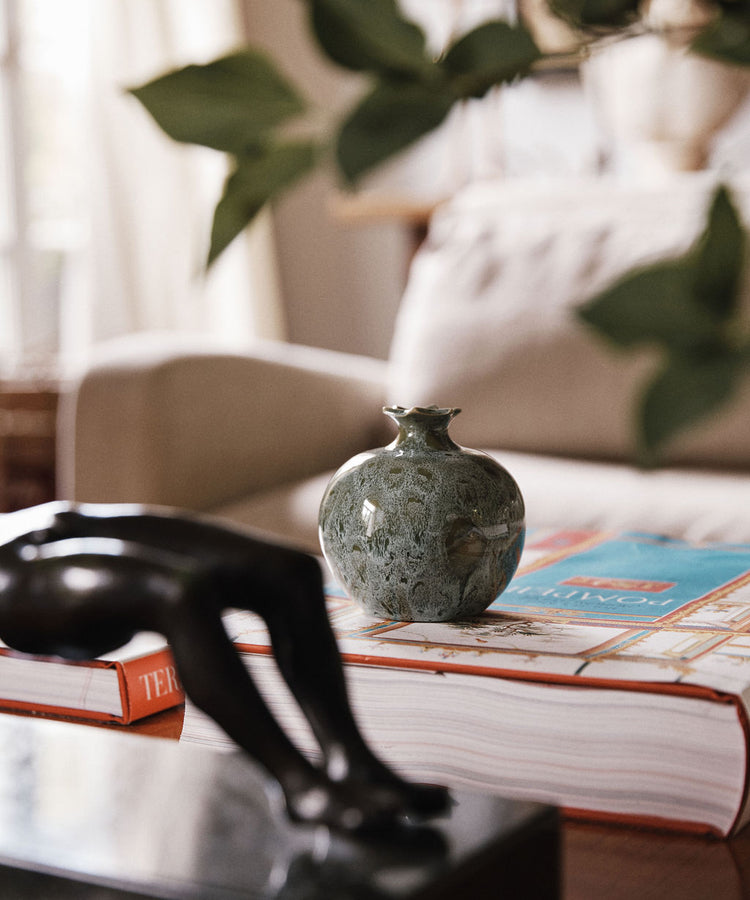 A Pom Bud Vase in green, inspired by handmade ceramic pomegranate designs from Granada ceramics, sits atop two stacked books on a table, with a blurred black sculpture in the foreground and a beige couch in the background.
