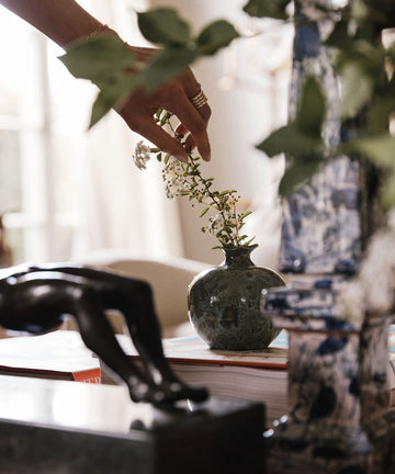 A hand places white flowers in the Pom Bud Vase on a table, surrounded by books, a bronze sculpture, a blue-and-white decor piece, and a handmade ceramic pomegranate inspired by Granada ceramics.