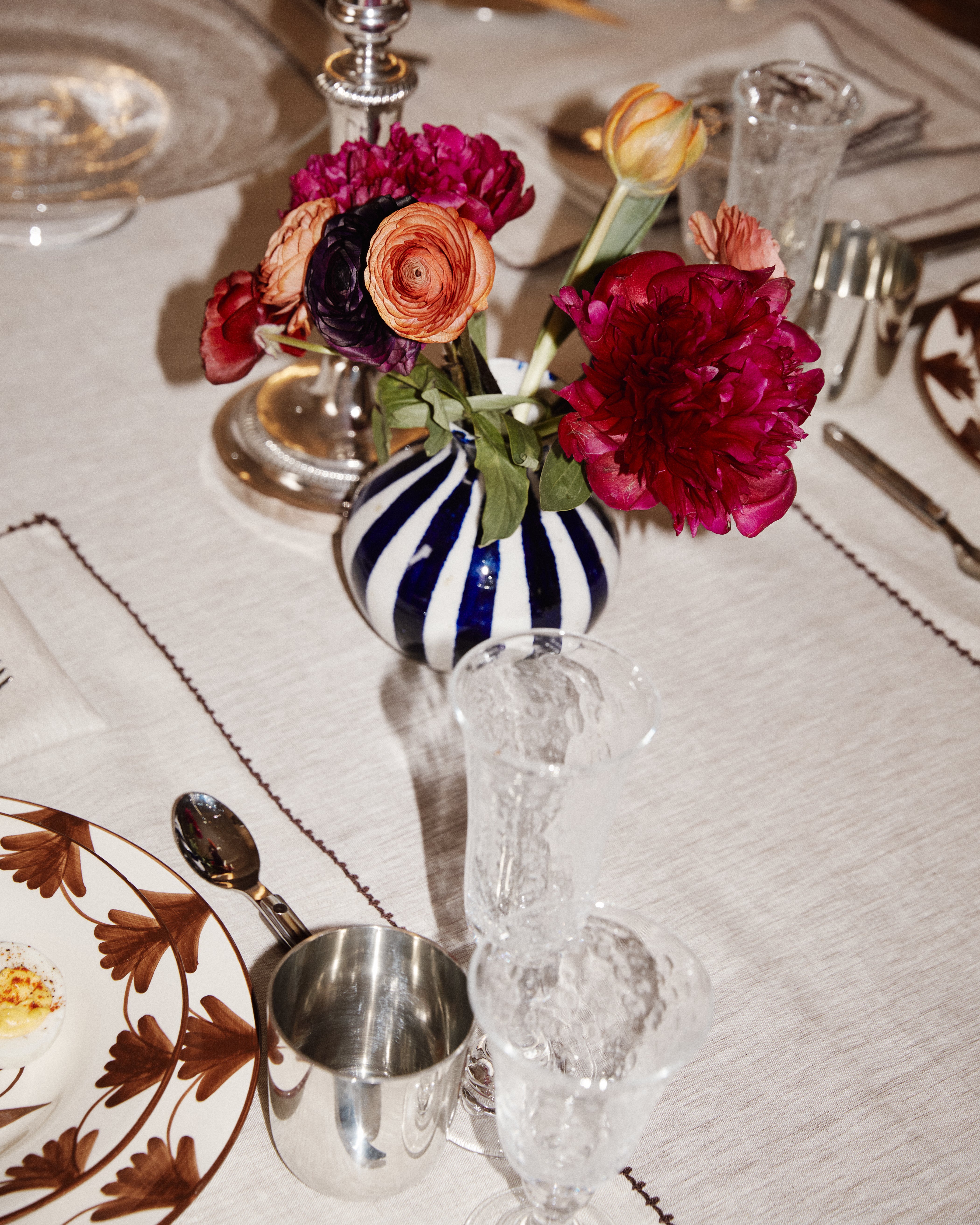 A Flora Bud Vase with cobalt stripes and pink and orange flowers sits on a set dining table with plates, glasses, and silverware.