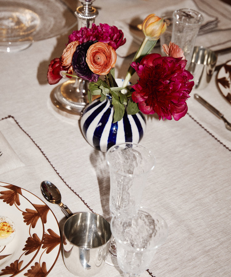 A Flora Bud Vase with cobalt stripes and pink and orange flowers sits on a set dining table with plates, glasses, and silverware.