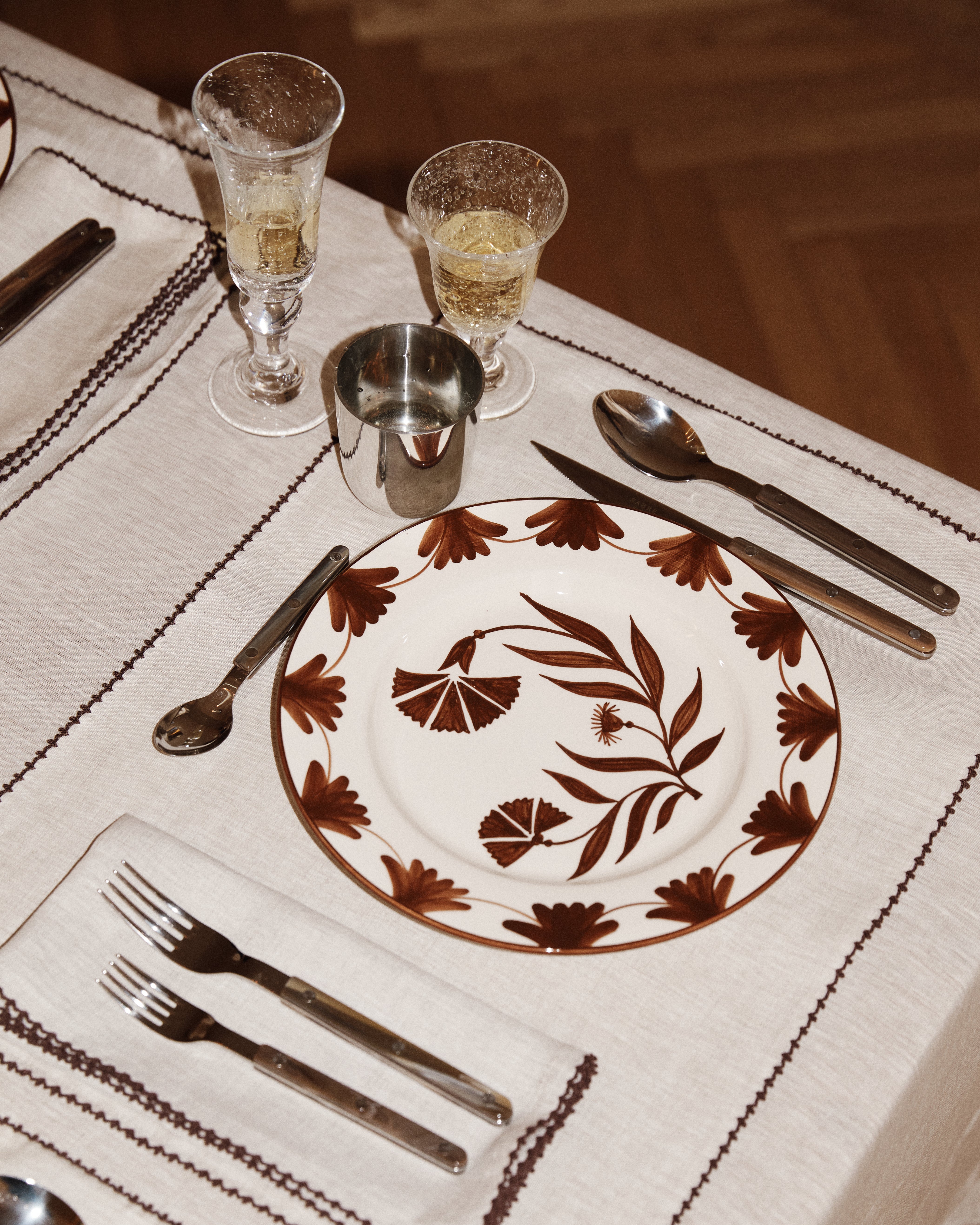 A dinner setting featuring a Catalina Dinner Plate, fork, knife, spoon, metal cup, glass with drink, and napkin on a beige tablecloth.