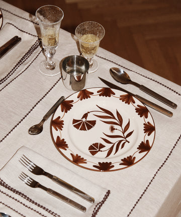 A dinner setting featuring a Catalina Dinner Plate, fork, knife, spoon, metal cup, glass with drink, and napkin on a beige tablecloth.