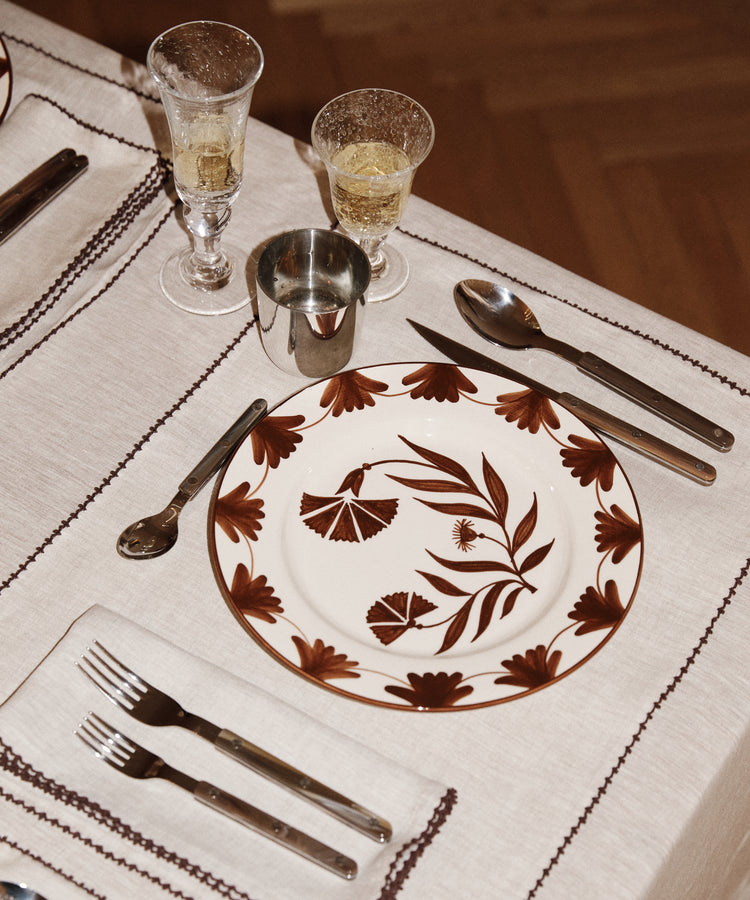 A dinner setting featuring a Catalina Dinner Plate, fork, knife, spoon, metal cup, glass with drink, and napkin on a beige tablecloth.