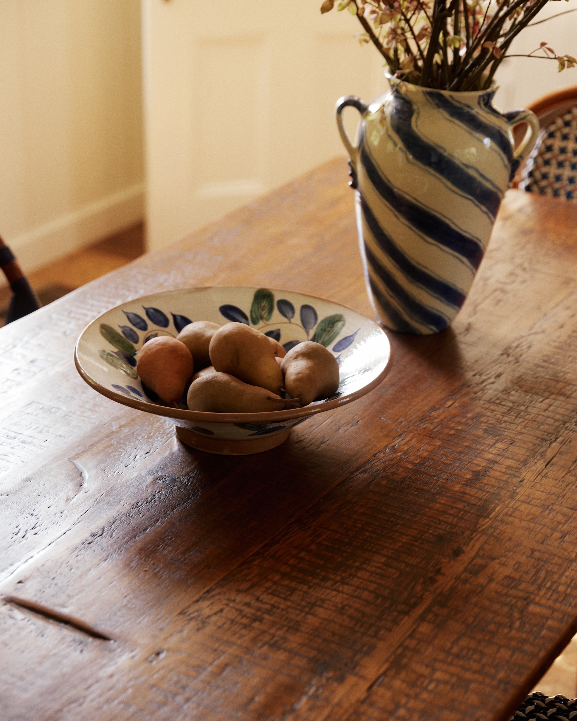 A Midnight Garden Serving Bowl filled with potatoes rests on a wooden table beside a blue-and-white striped vase with dried flowers.