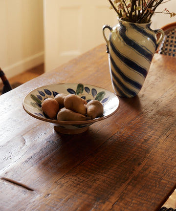 A Midnight Garden Serving Bowl filled with potatoes rests on a wooden table beside a blue-and-white striped vase with dried flowers.