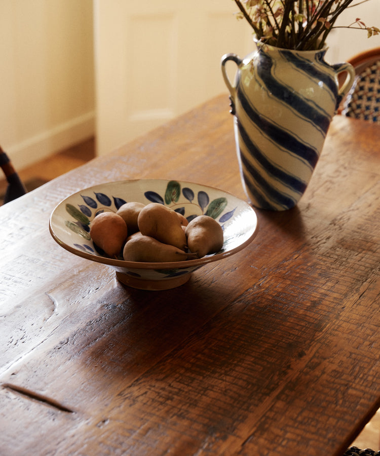A Midnight Garden Serving Bowl filled with potatoes rests on a wooden table beside a blue-and-white striped vase with dried flowers.