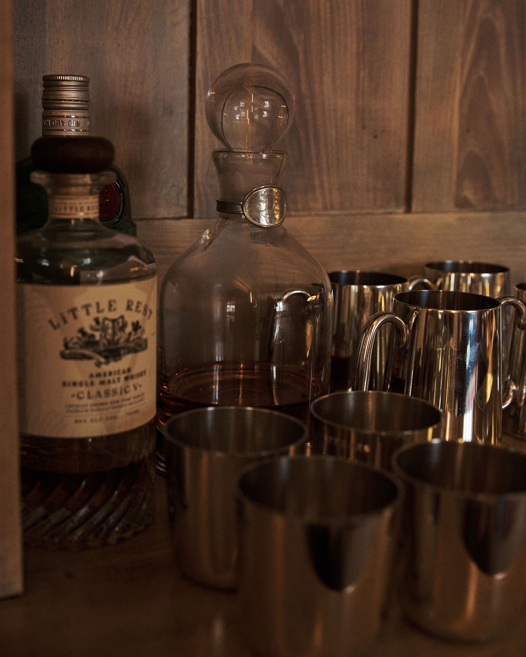 The Alec Decanter, featuring a hand-finished pewter collar, sits beside metal cups on a wooden surface against a wood-paneled wall.