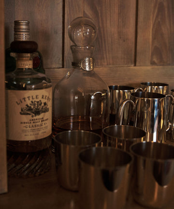 The Alec Decanter, featuring a hand-finished pewter collar, sits beside metal cups on a wooden surface against a wood-paneled wall.