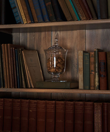 A Bonbon Jar filled with cookies sits on a shelf among old books in a wooden bookcase.