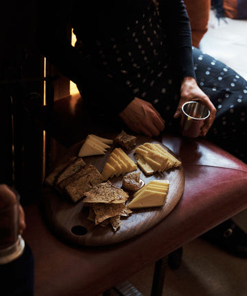 A person in a dark, polka-dotted outfit sits holding a metal cup beside an Alden Egg Board with assorted cheeses and crackers on a leather bench.