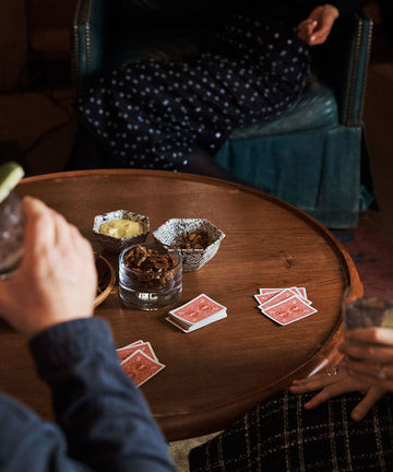 Three people sit around a wooden table with playing cards and drinks in hand, enjoying snacks served in the handcrafted Celine Snack Bowl.