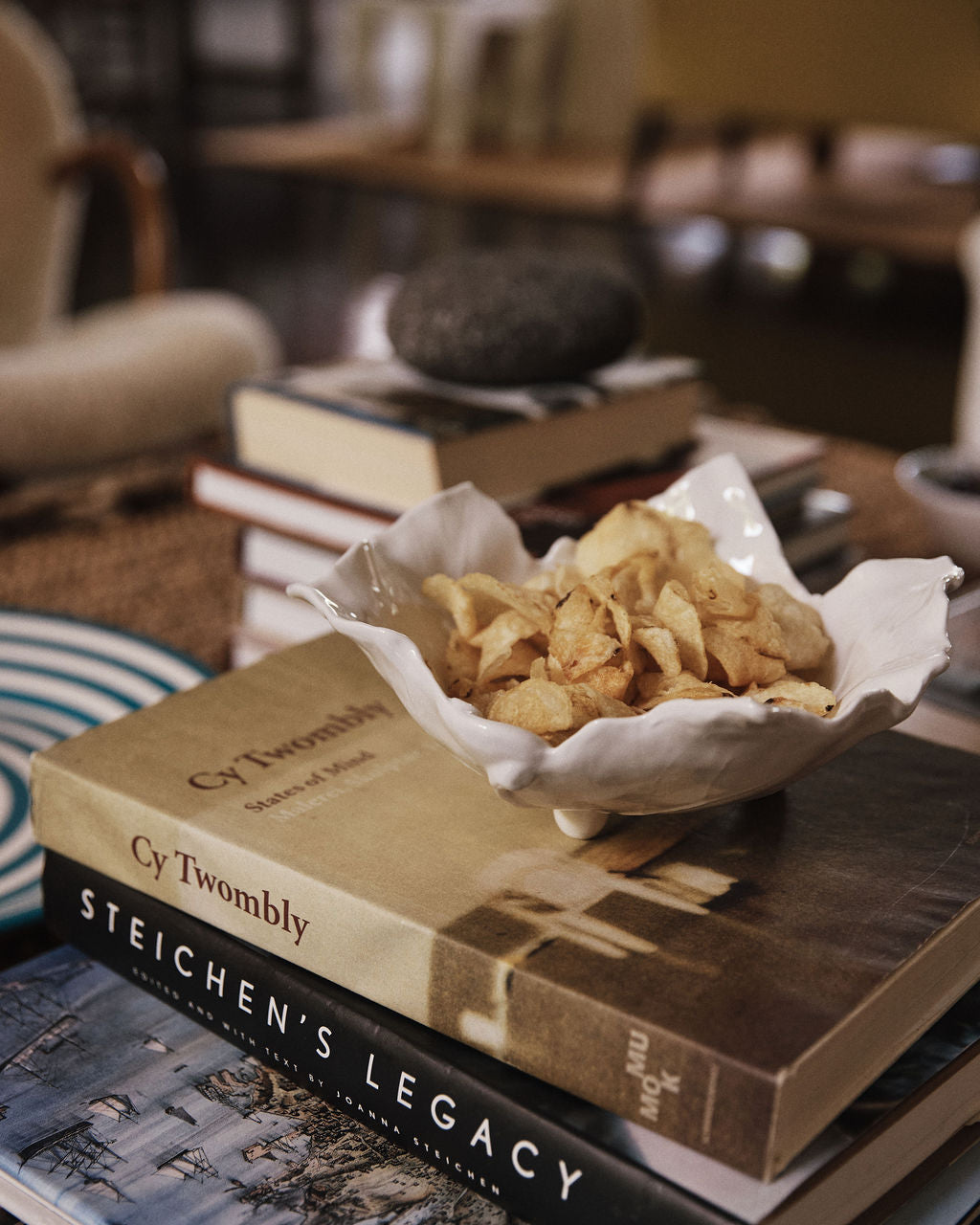 A Leaf Serving Bowl filled with potato chips sits atop art books, including Cy Twombly and Steichen's Legacy, on a table decorated with other books and decor items.
