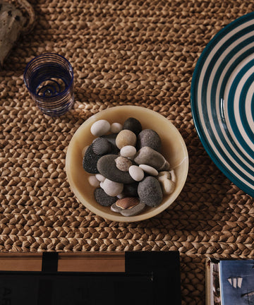 The Akila Decorative Bowl, handcrafted from yellow Egyptian alabaster and filled with smooth stones and seashells, sits on a woven surface beside a blue-striped plate and blue-rimmed glass, capturing the artistry of Luxor.