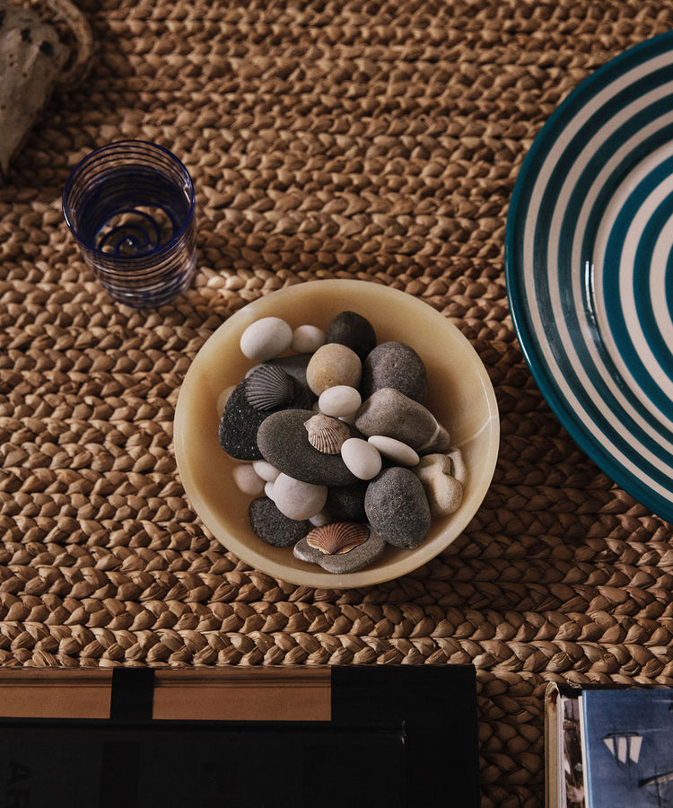 The Akila Decorative Bowl, handcrafted from yellow Egyptian alabaster and filled with smooth stones and seashells, sits on a woven surface beside a blue-striped plate and blue-rimmed glass, capturing the artistry of Luxor.