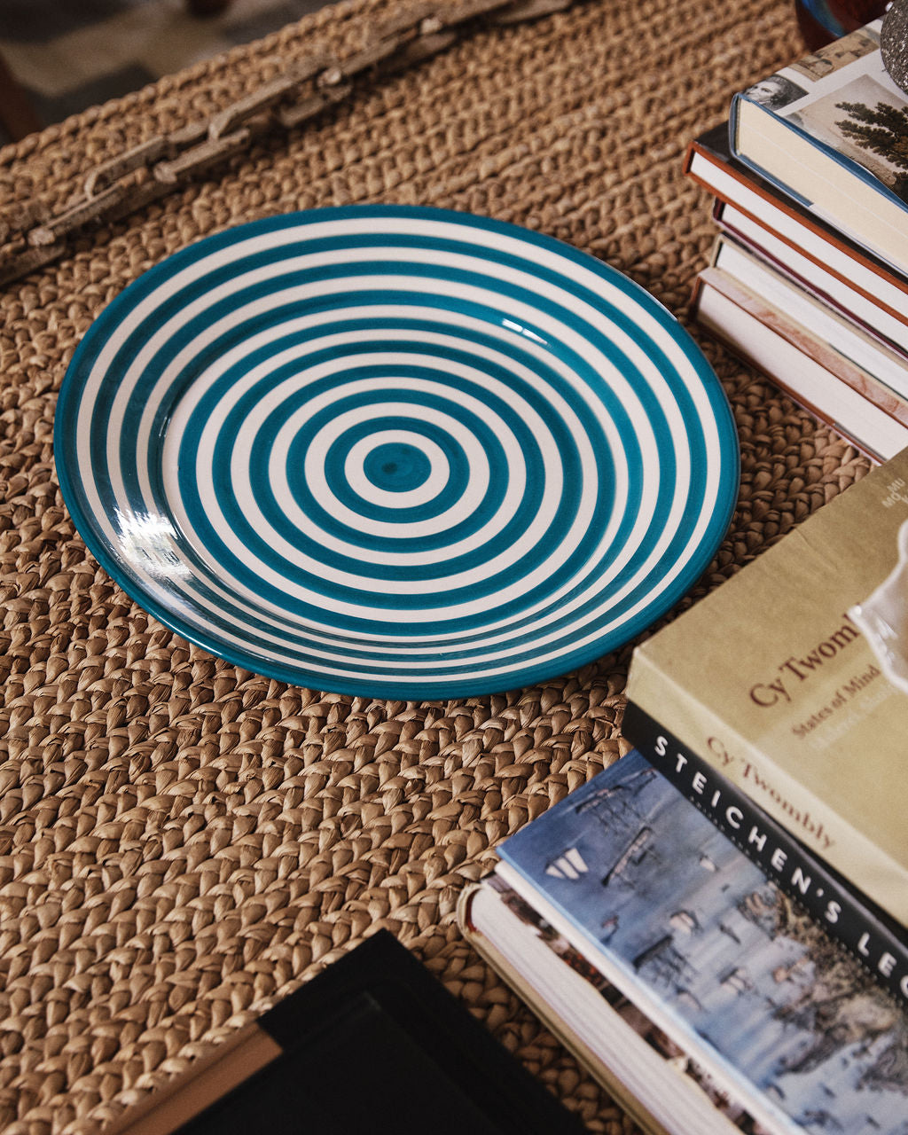 A unique Natalia Platter, featuring blue and white concentric stripes, sits on a woven mat surrounded by stacks of books.