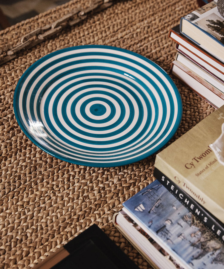 A unique Natalia Platter, featuring blue and white concentric stripes, sits on a woven mat surrounded by stacks of books.