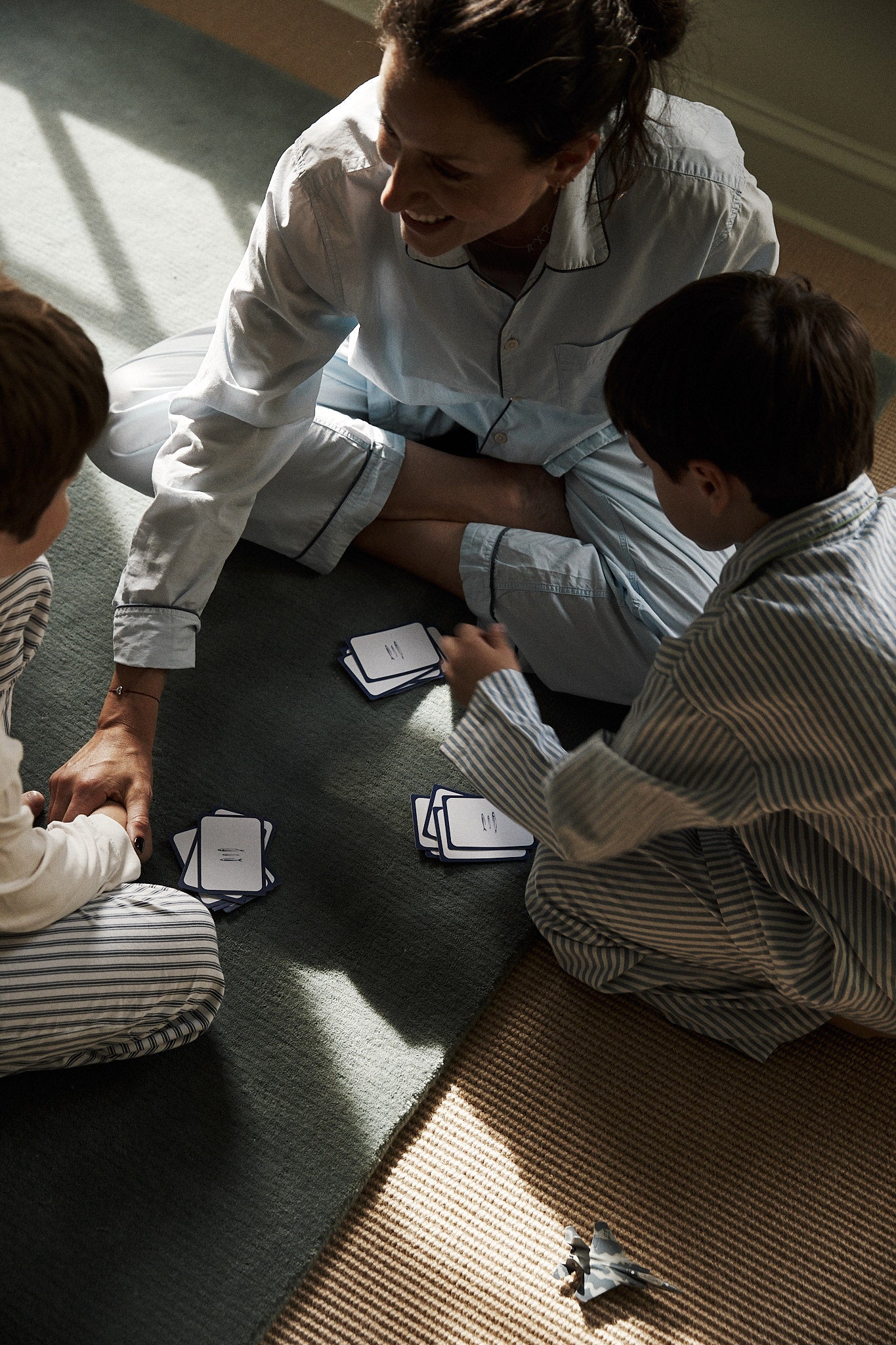 An adult and two children in pajamas sit on the floor playing a card game together with PORTA Playing Cards, while a toy airplane rests nearby.