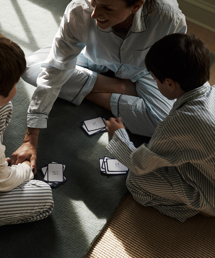 An adult and two children in pajamas sit on the floor playing a card game together with PORTA Playing Cards, while a toy airplane rests nearby.