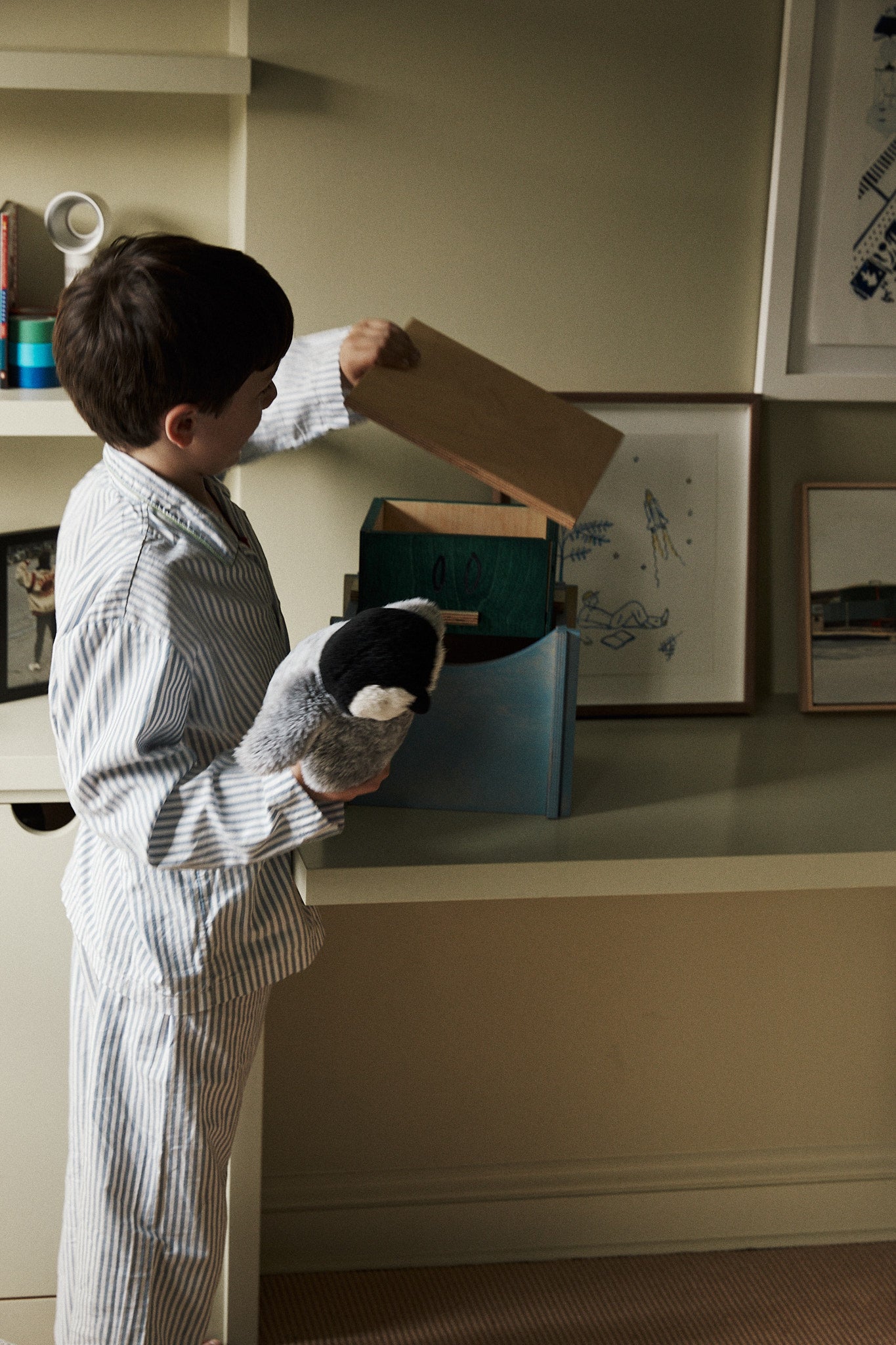 A young boy in striped pajamas holds a stuffed penguin and looks into the Milo Keepsake Box by House of RoRo on a shelf in a softly lit room.
