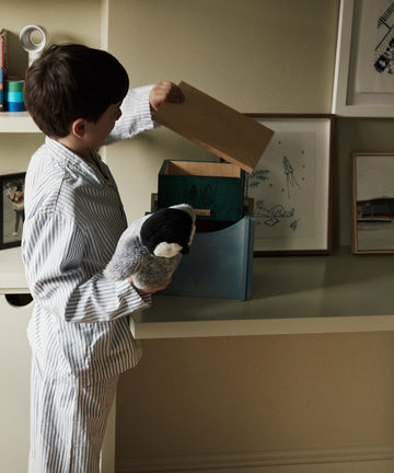 A young boy in striped pajamas holds a stuffed penguin and looks into the Milo Keepsake Box by House of RoRo on a shelf in a softly lit room.