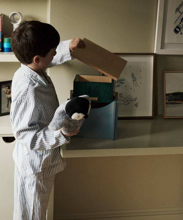 A young boy in striped pajamas holds a stuffed penguin and looks into the Milo Keepsake Box by House of RoRo on a shelf in a softly lit room.