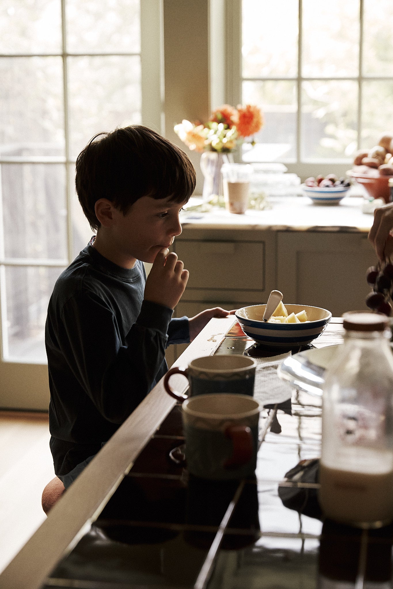 Child sitting at a kitchen table with a bowl of fruit and a cup, with a window in the background.