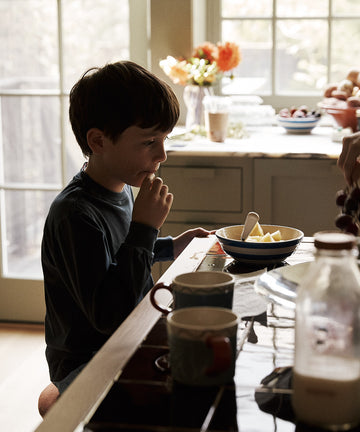 Child sitting at a kitchen table with a bowl of fruit and a cup, with a window in the background.