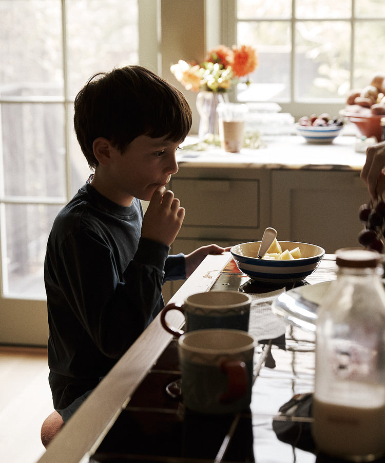 Child sitting at a kitchen table with a bowl of fruit and a cup, with a window in the background.