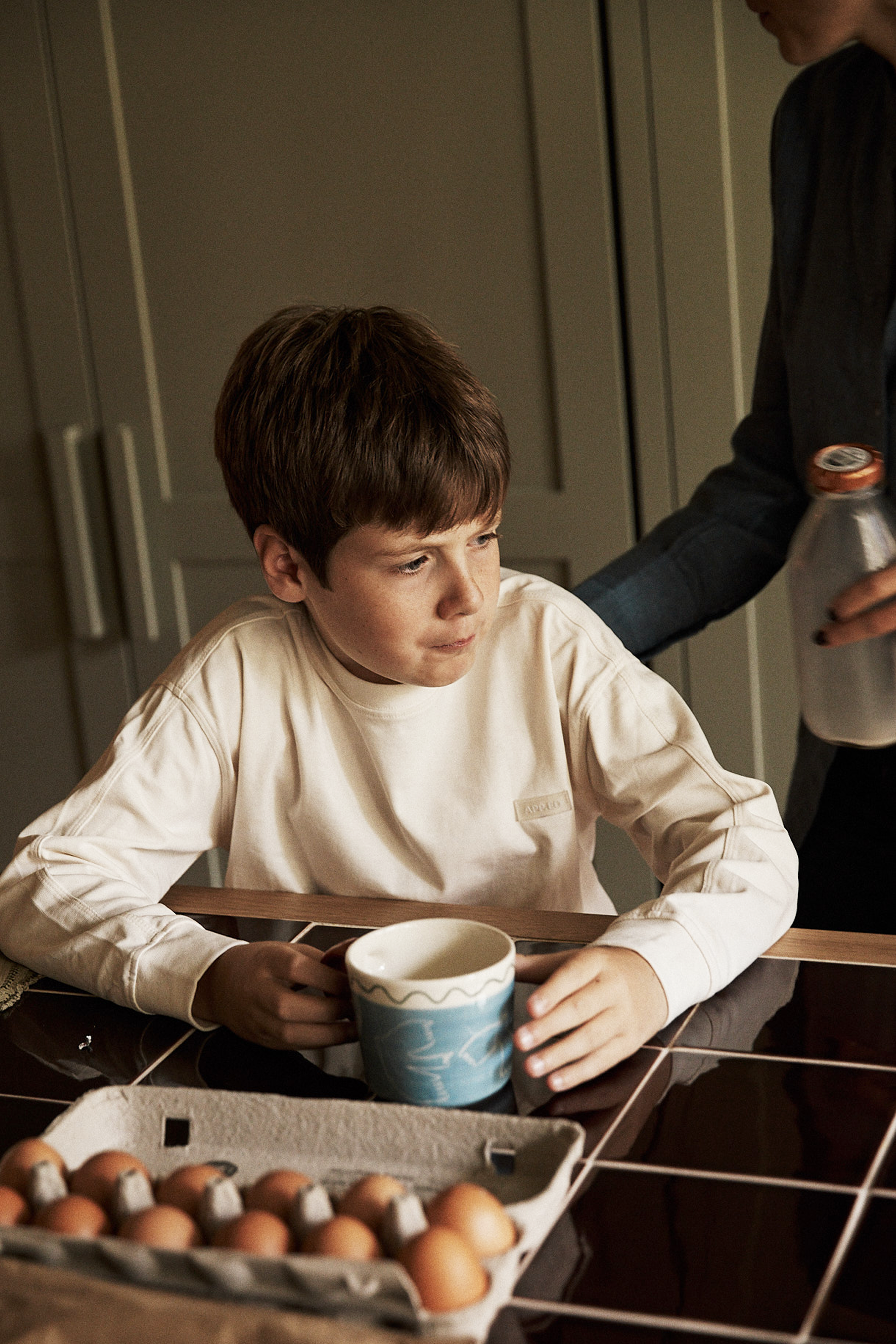 A boy sits at a kitchen table holding a Colombe Mug, with a carton of eggs in front of him, as an adult hands him a bottle of milk.
