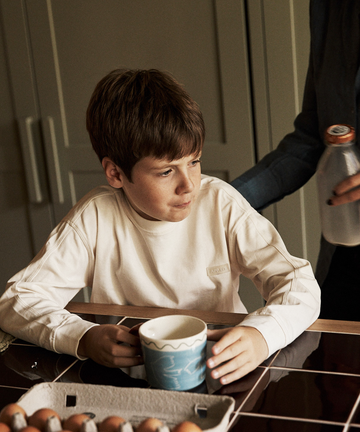 A boy sits at a kitchen table holding a Colombe Mug, with a carton of eggs in front of him, as an adult hands him a bottle of milk.