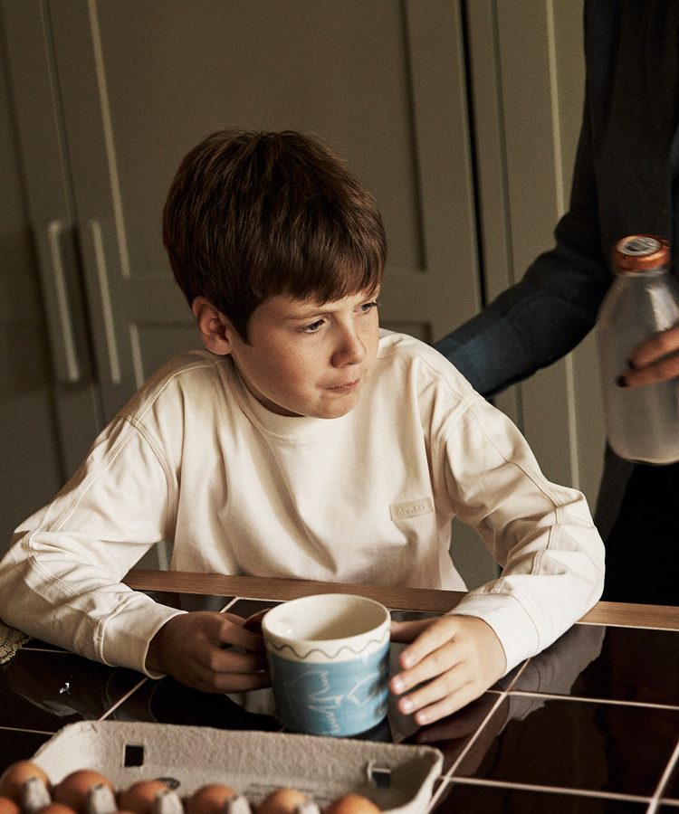 A boy sits at a kitchen table holding a Colombe Mug, with a carton of eggs in front of him, as an adult hands him a bottle of milk.