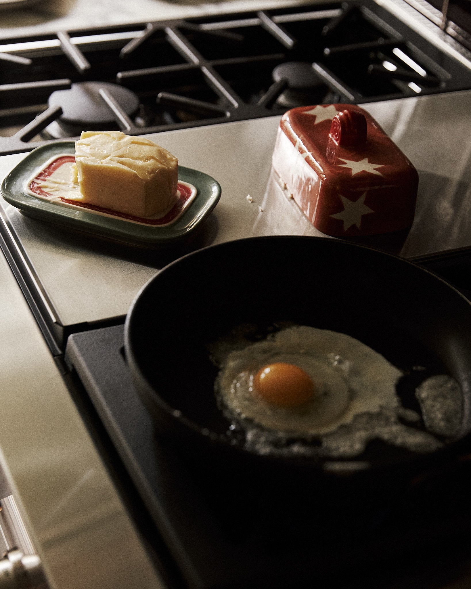 A raw egg fries in a black pan on the stove, while a Butterfingers Butter Dish with butter rests on the counter nearby.