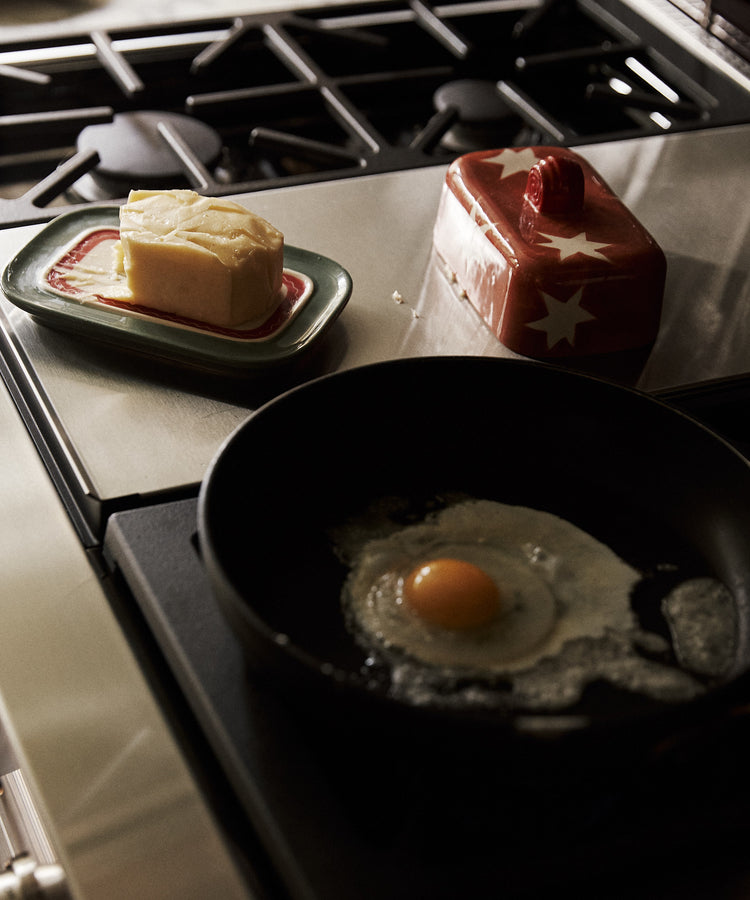 A raw egg fries in a black pan on the stove, while a Butterfingers Butter Dish with butter rests on the counter nearby.