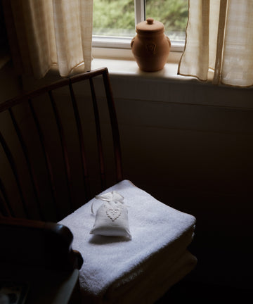 A stack of white towels with a Lavender Sachet handmade in Portugal rests on a chair by a window with sheer curtains and a ceramic jar on the sill.