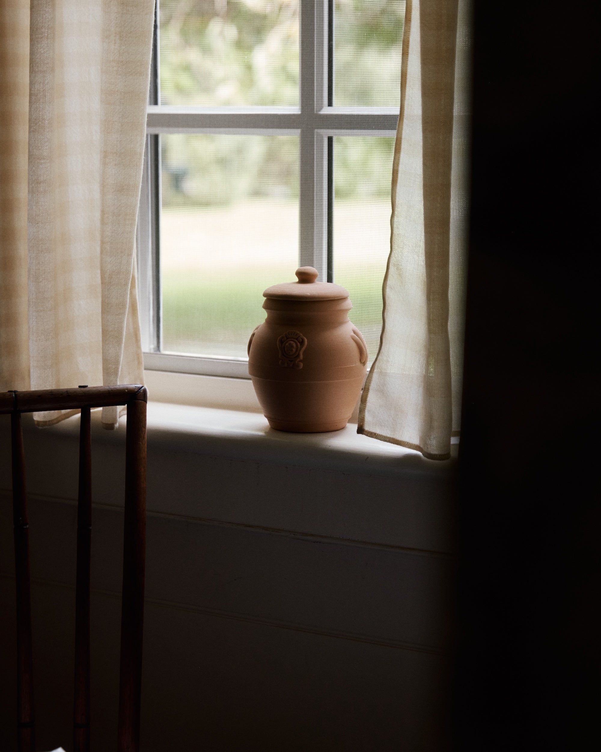 The Pot Pourri in Terracotta Jar with a lid sits on a windowsill between light curtains, bathed in soft daylight through the window.
