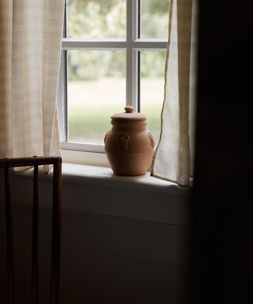 The Pot Pourri in Terracotta Jar with a lid sits on a windowsill between light curtains, bathed in soft daylight through the window.