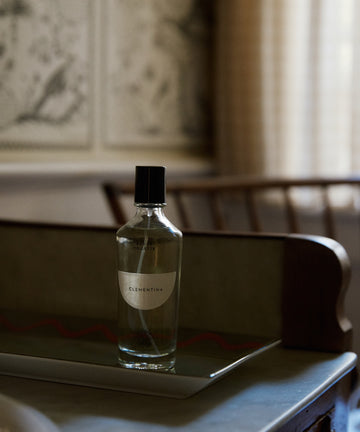 A clear glass bottle of Clementina Eau de Toilette rests on a tray atop a table, capturing bergamot notes, with a blurred wooden chair and patterned wall behind.
